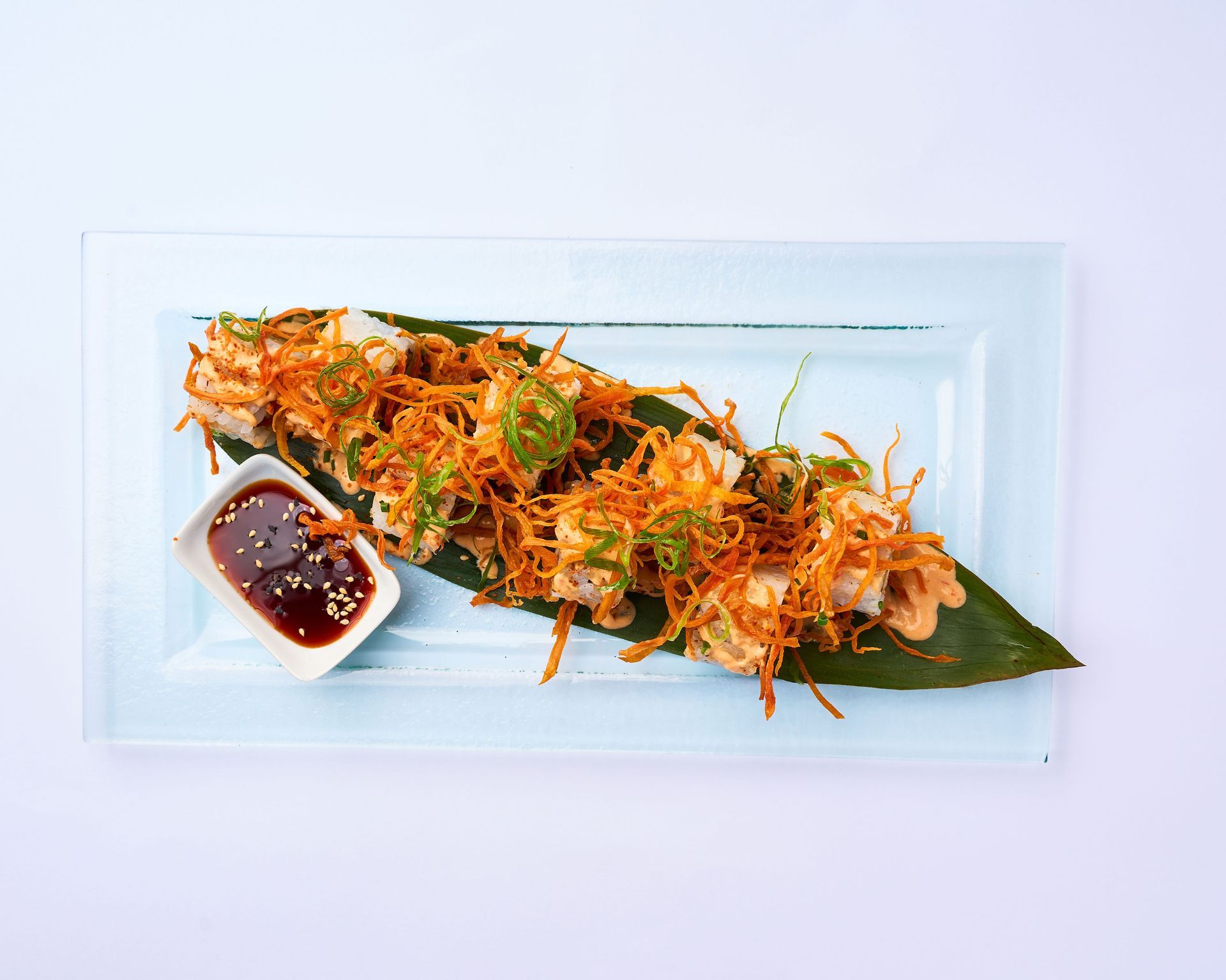 Un plato rectangular de cristal contiene sushi cubierto con zanahorias ralladas crujientes, cebolletas y una guarnición de salsa de soja para mojar.