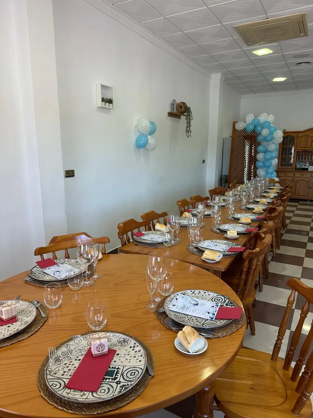 Mesa larga puesta para una comida. Platos, vasos y pan están dispuestos. Globos azules y blancos decoran la pared.