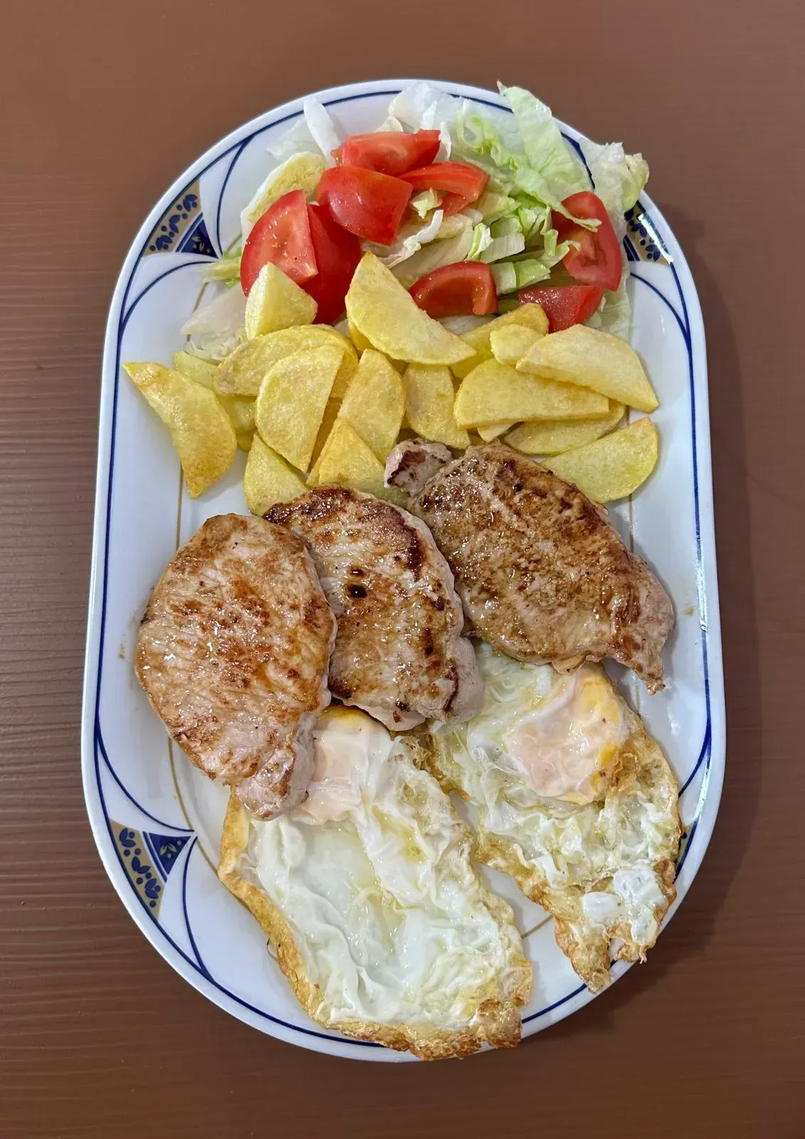 Plato de comida: cerdo, huevos fritos, gajos de patata y ensalada de lechuga y tomate.