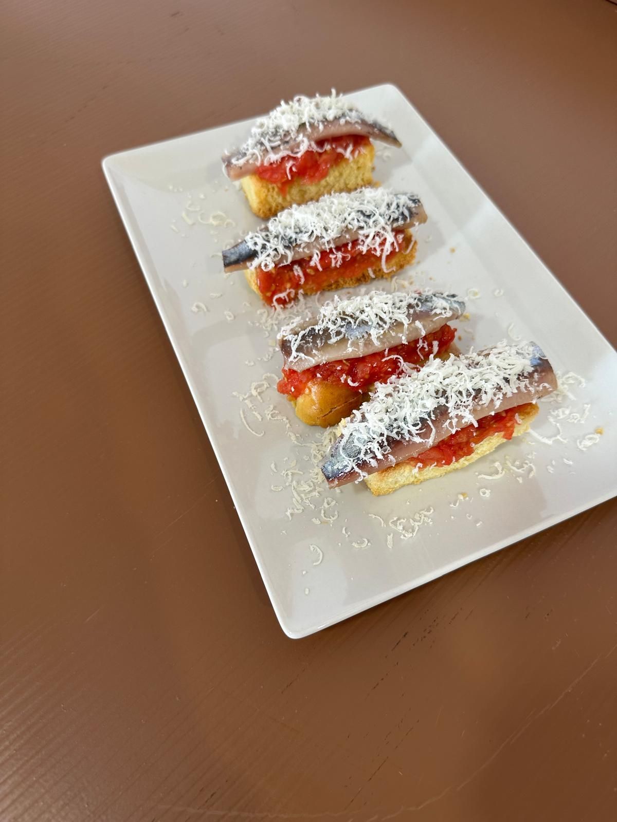 Cinco tapas con anchoas, salsa de tomate y queso rallado en un plato blanco, sobre una superficie marrón.