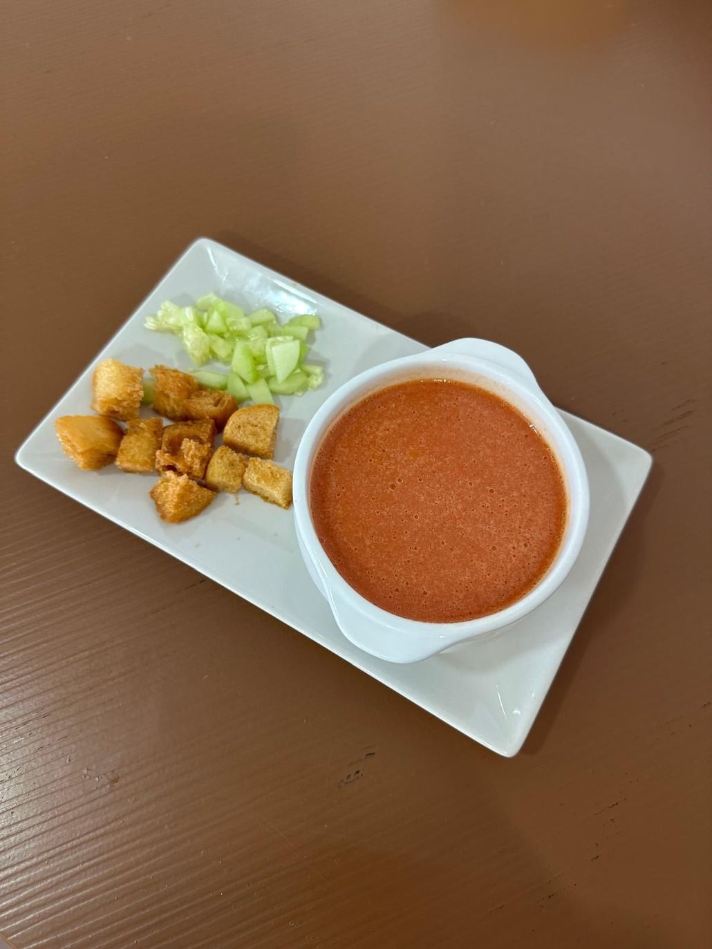 Plato blanco con un tazón de sopa roja, cubos de tofu y pepino.