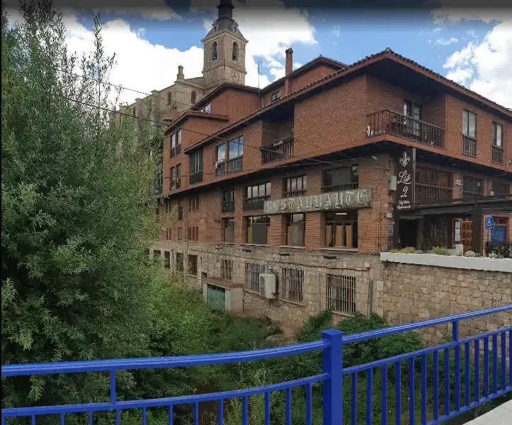 Edificio de ladrillo con balcones y base de piedra, junto a un río con barandilla azul. Torre de la iglesia al fondo.