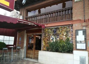Fachada de restaurante con toldo rojo, puerta de madera, exterior de ladrillo, balcón arriba y asientos al aire libre.