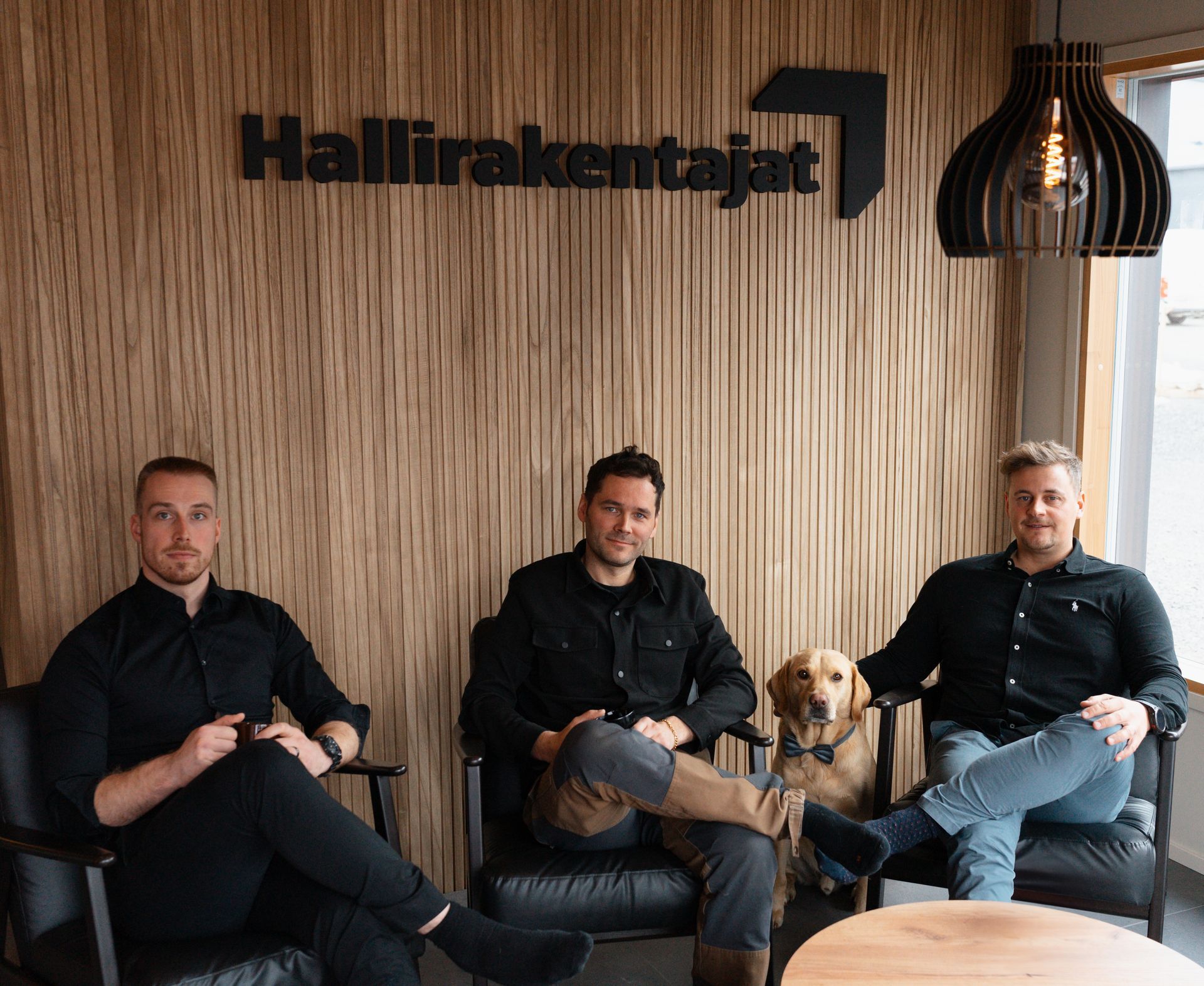 Kolme miestä ja koira istuvat kyltin edessä, jossa lukee hallirakentajat