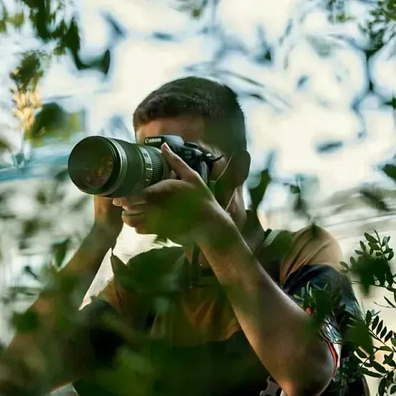 Persona tomando una fotografía con una cámara; rodeada de follaje verde.