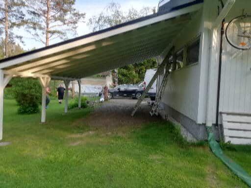 Taloon kiinnitetty autokatos. Valkoinen katto ja tuet, vihreä nurmikko.