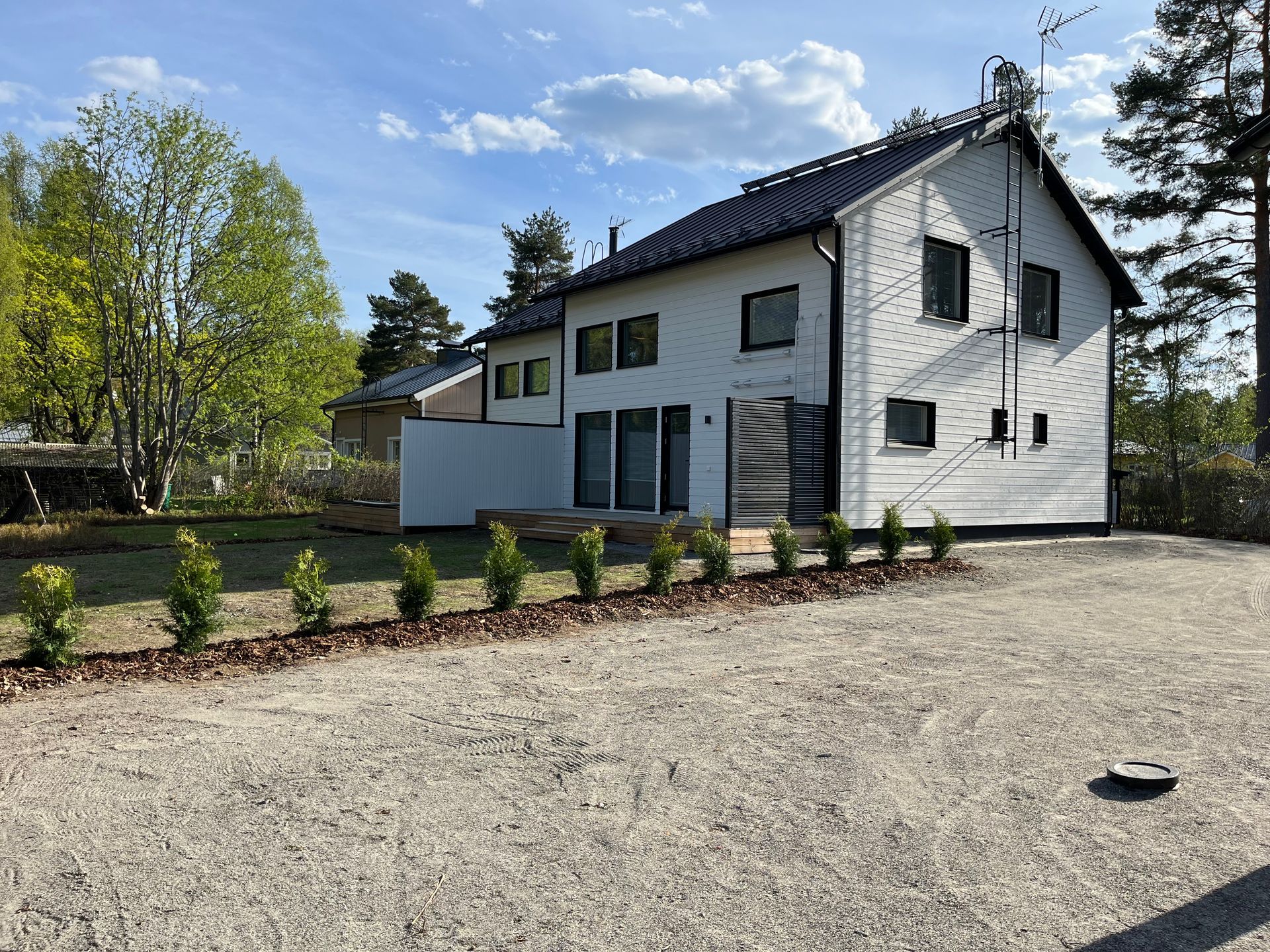 Valkoinen kaksikerroksinen talo tummalla katolla, soran, pienten puiden ja vehreyden ympäröimänä pilvisen taivaan alla.