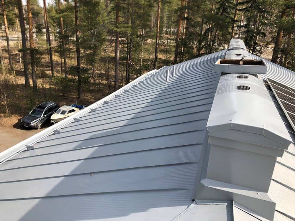 Harmaa peltikatto tuuletusaukoilla, näkymä metsään. Autoja pysäköitynä alapuolelle.