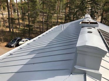 Hopeanvärinen metallinen katto tuuletusaukoilla ja aurinkopaneeleilla. Autot pysäköitynä puiden lähelle.