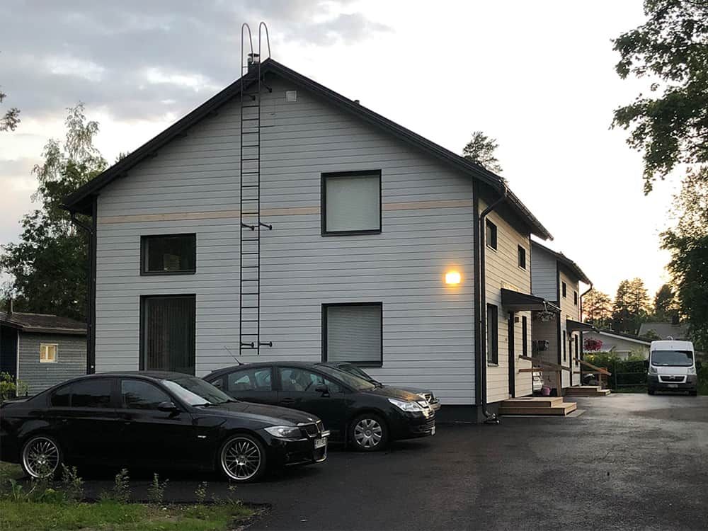 Kaksikerroksinen valkoinen rakennus, jonka edessä on pysäköitynä autoja; musta BMW ja hopeanvärinen auto. Sivussa tikkaat.