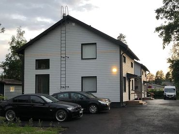 Kaksikerroksinen valkoinen rakennus tummalla katolla, autoja pysäköitynä edessä. Tikkaat sivussa.