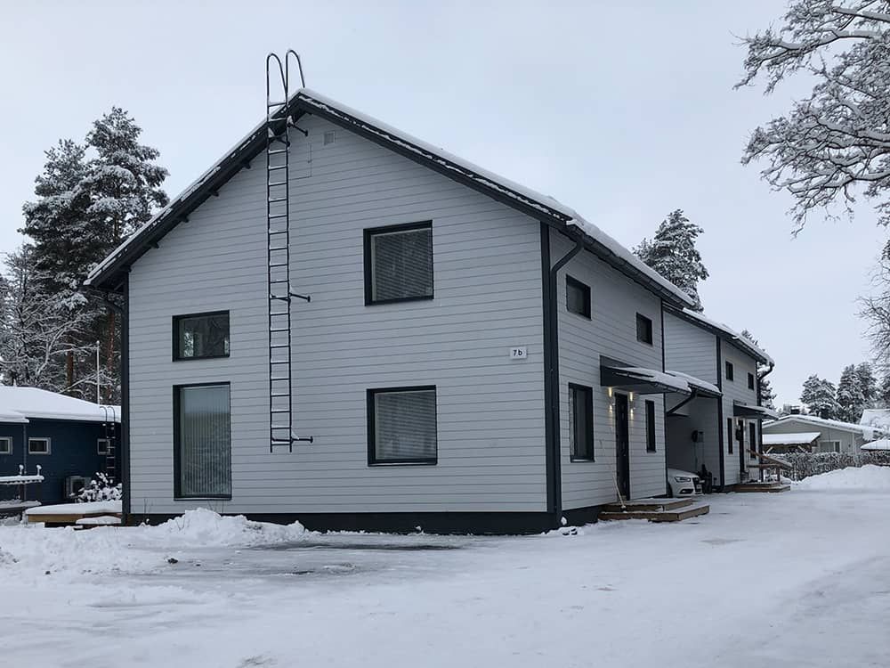 Kaksikerroksinen vaaleanharmaa rakennus tummilla reunuksilla, lumen peittämä maa, tikkaat sivulla, talvinen ympäristö.