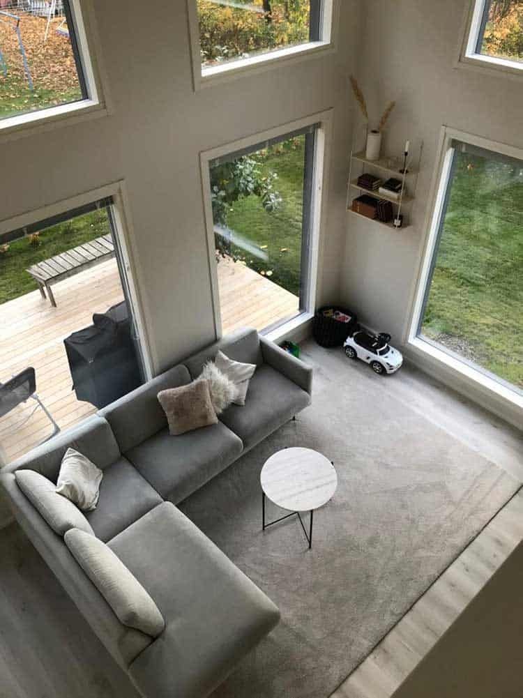 Ylhäältä katsottuna moderni olohuone, jossa on harmaa sohvaryhmä, pyöreä pöytä, suuret ikkunat ja ulkonäkymä.