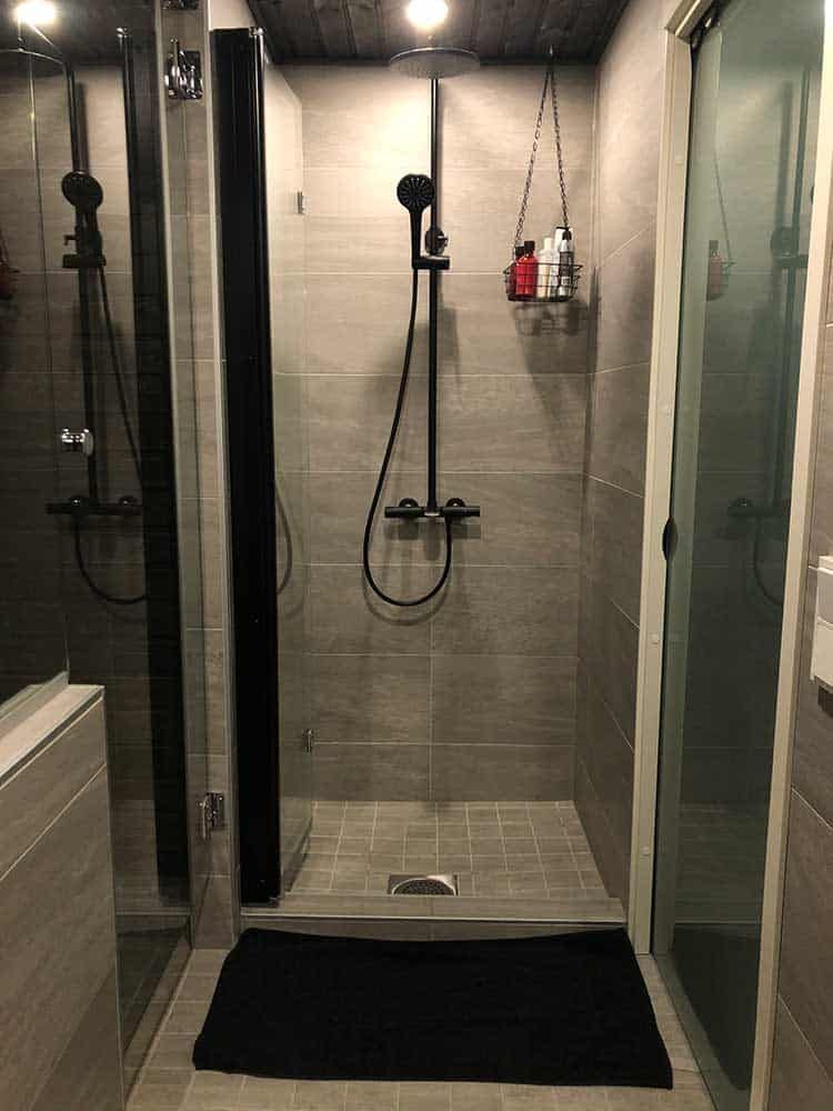 Moderni suihku, jossa on harmaat laatat, musta suihkupää ja musta matto.