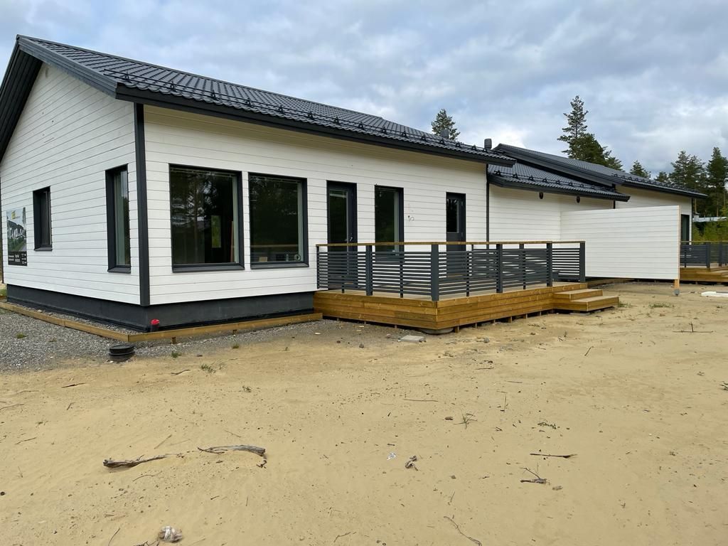 Valkoinen talo tummilla reunoilla, mustalla katolla, puisella terassilla ja pilvisellä taivaalla.