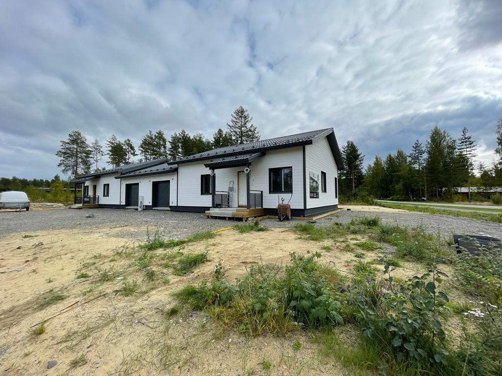 Valkoinen talo mustilla reunuksilla, sorapiha, pilvinen taivas, puiden ympäröimänä.