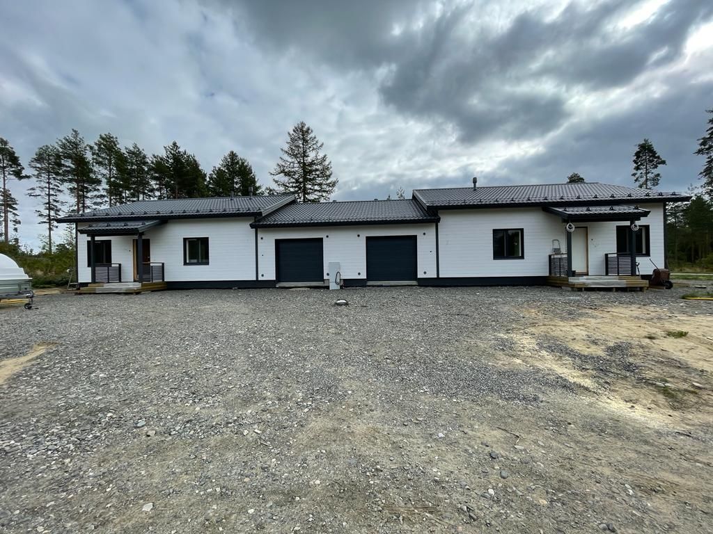 Valkoinen talo, jossa mustat autotallin ovet, ikkunat ja listat, sijaitsee sora-alueella metsän taustalla.