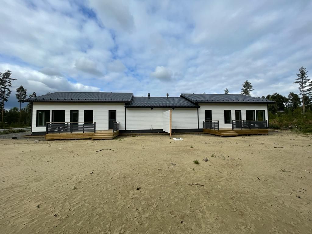 Valkoinen talo tummalla katolla ja puisilla terasseilla hiekkaisella tontilla pilvisen taivaan alla.