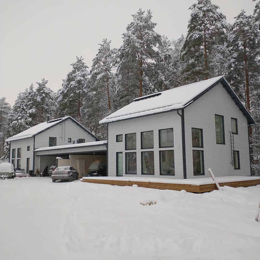 Kaksi vaaleanharmaata taloa lumisessa metsässä, katetulla autokatoksella.