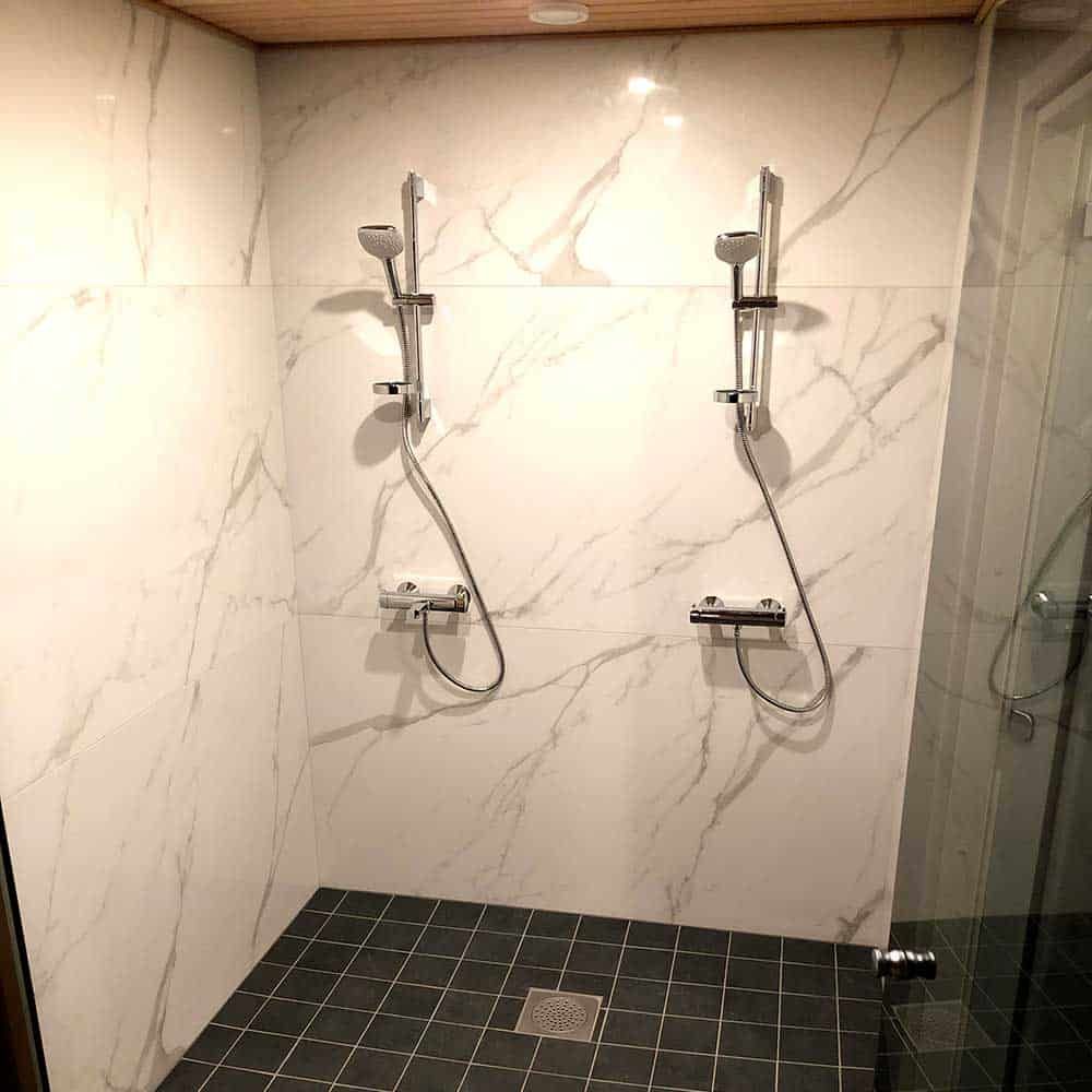 Kaksi suihkua kromivalaisimilla, valkoisilla marmoriseinillä ja mustalla laattalattialla.