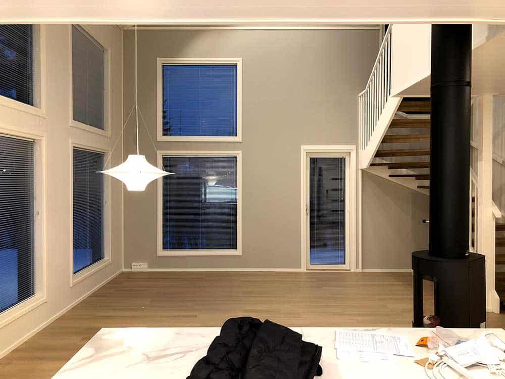 Moderni sisustus, jossa vaalea puulattia ja harmaat seinät. Ikkunat, portaikko ja musta takka.