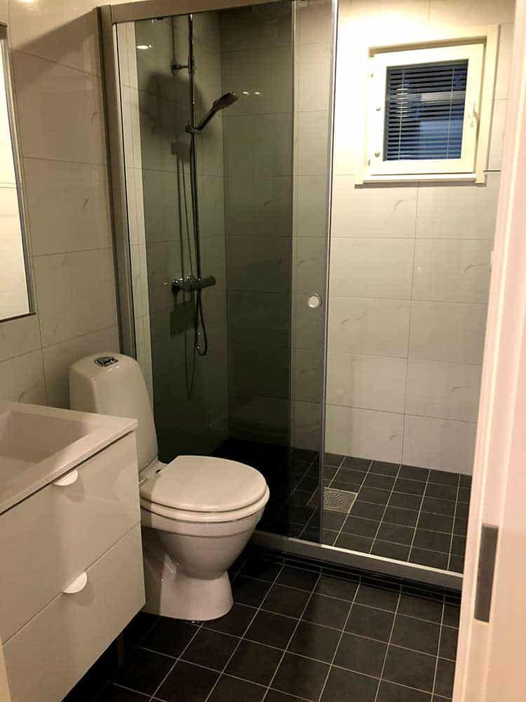 Pieni kylpyhuone, jossa wc, suihku, pesuallas ja ikkuna. Harmaat ja mustat laatat kaikkialla.