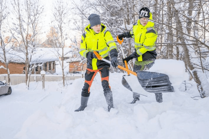 Kaksi keltaisiin turvavarusteisiin pukeutunutta ihmistä lapioi lunta lumisella asuinalueella.