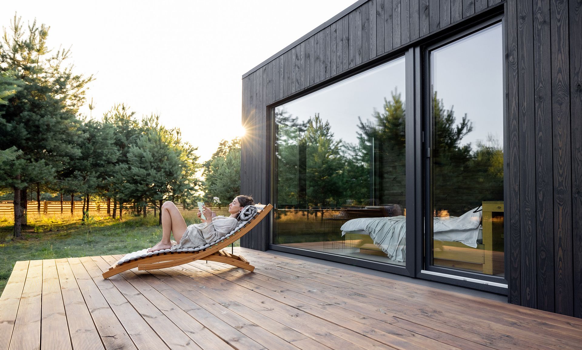 Henkilö rentoutuu puisella terassilla modernin mökin vieressä, auringonlasku puiden takana.