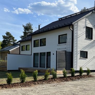Moderni valkoinen talo mustilla yksityiskohdilla ja puisella terassilla, sininen taivas yläpuolella.