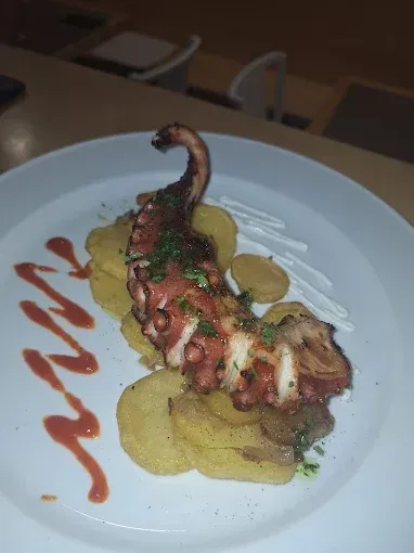 Tentáculo de pulpo a la parrilla sobre patatas fritas, rociado con salsas, servido en un plato blanco.