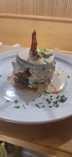 Una pila de comida con salsa en un plato blanco, adornada con verduras. Un trozo de comida marrón encima.