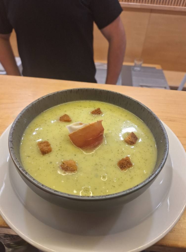 Sopa cremosa verde en un recipiente gris, adornada con crutones y una loncha de jamón.