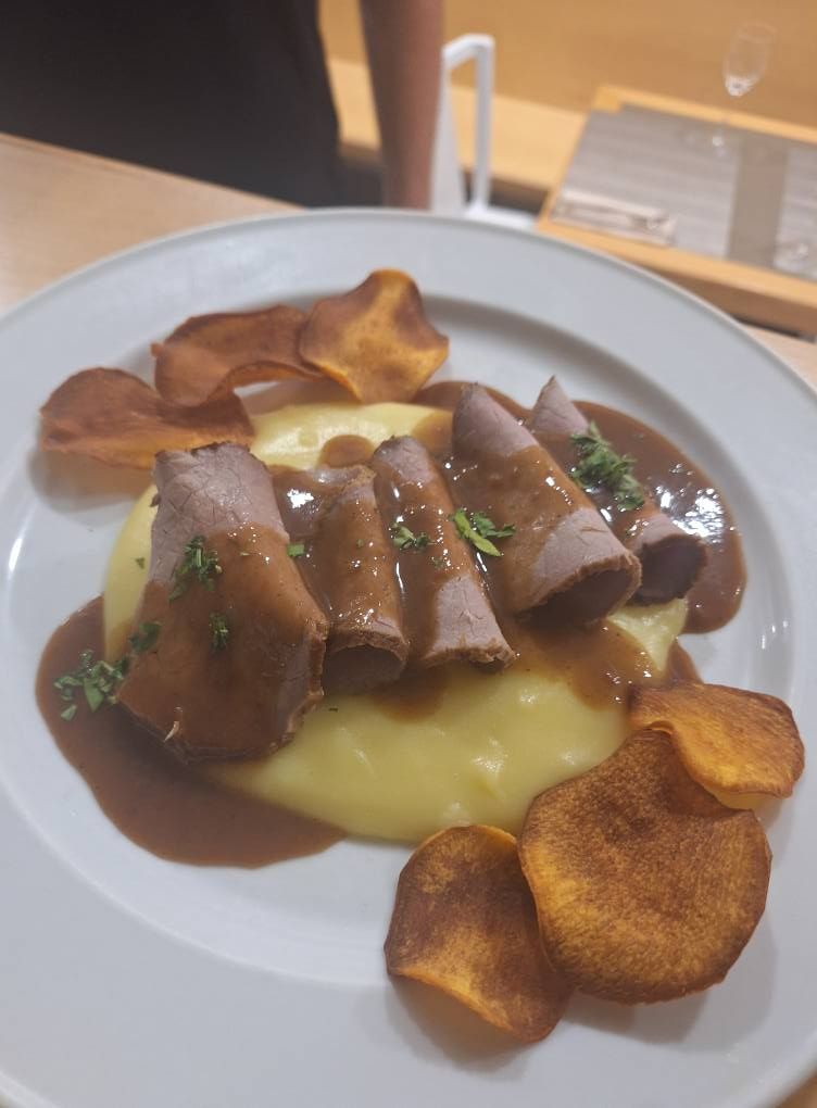 Carne en rodajas con salsa sobre puré de papas, adornada con hierbas y chips de batata en un plato blanco.