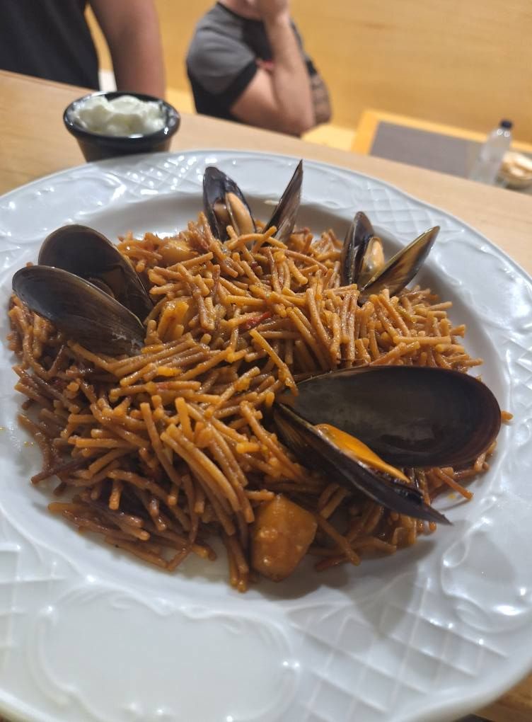 Fideuá con mejillones en un plato blanco. Al fondo, una persona y un plato pequeño con salsa.