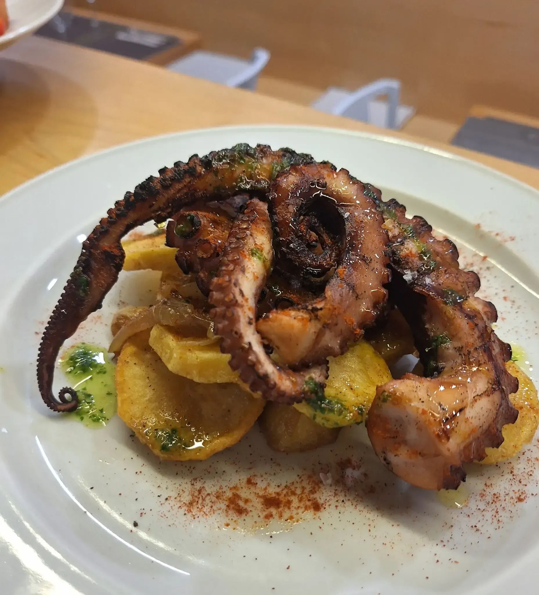Tentáculos de pulpo a la parrilla con patatas, rociados con salsa y espolvoreados con pimentón, servidos en un plato blanco.