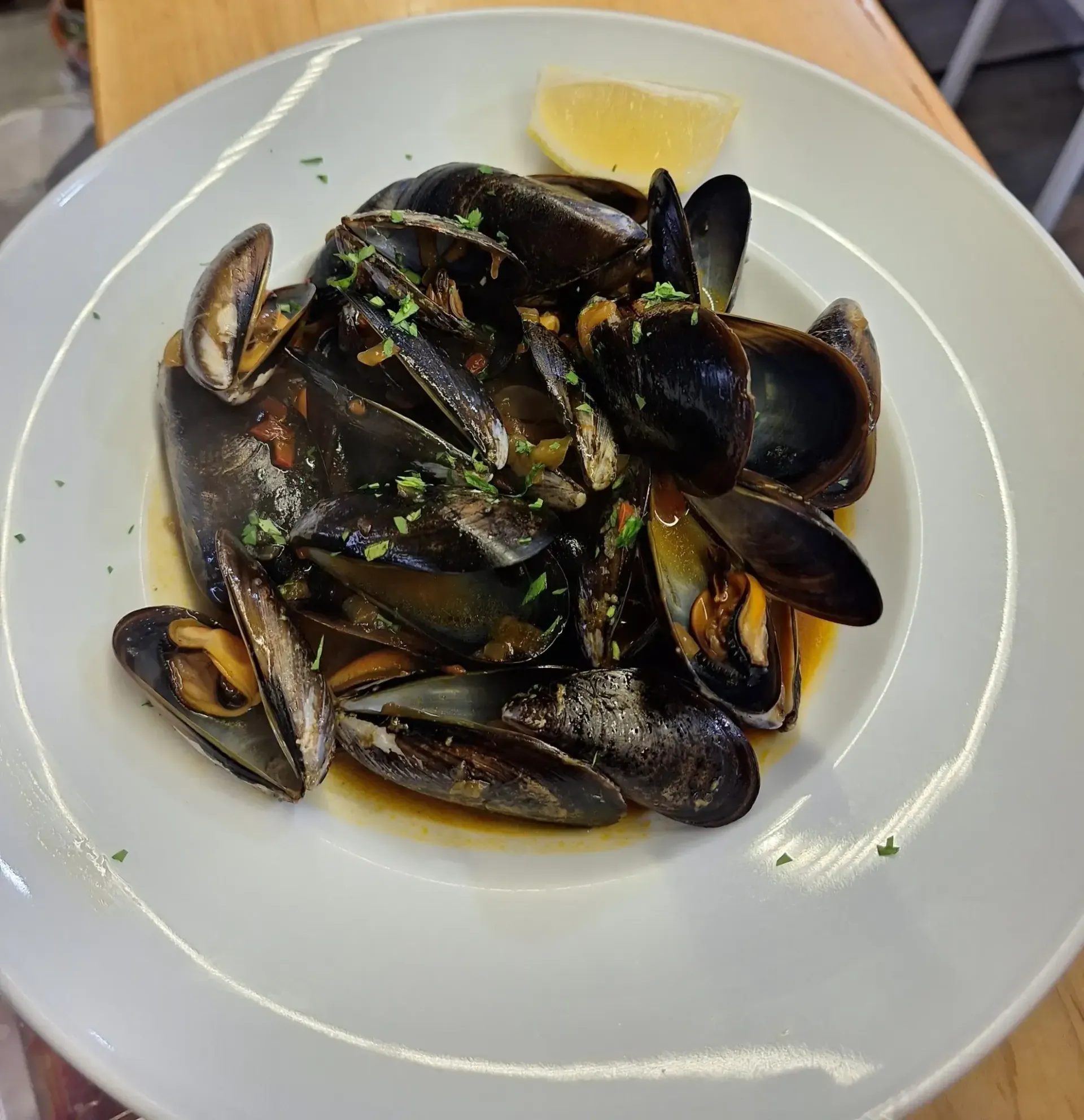 Mejillones al vapor en salsa oscura en plato blanco con rodaja de limón.