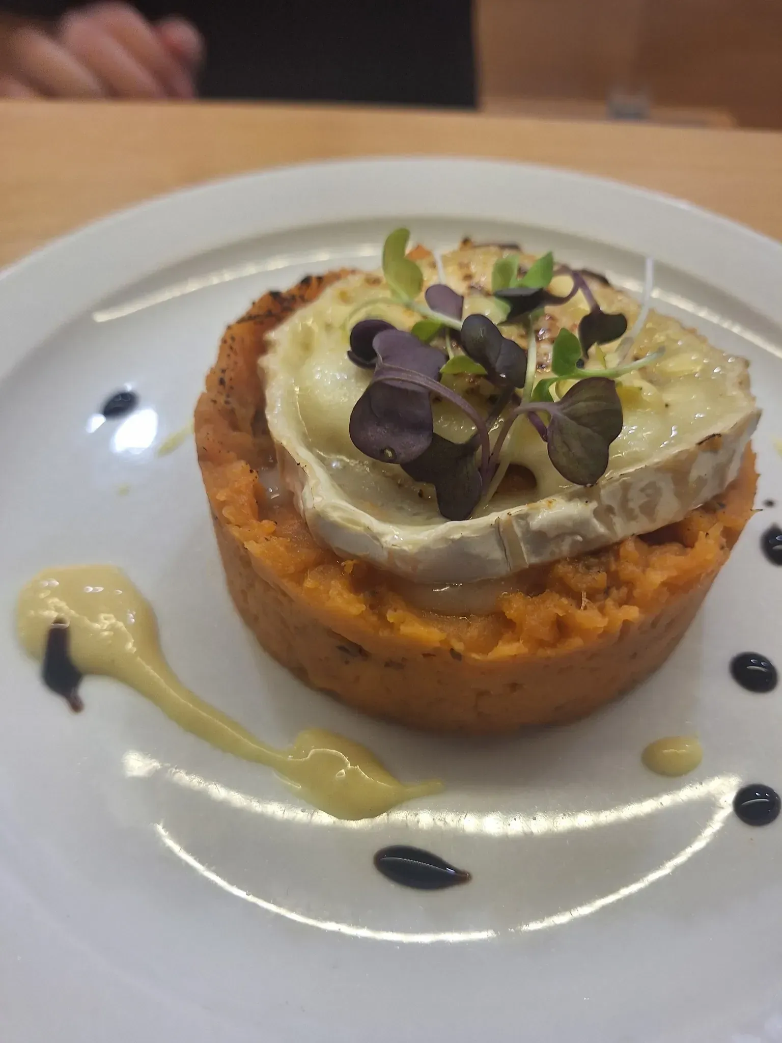 Una tarta salada pequeña y redonda cubierta con queso derretido y brotes, rociada con salsa en un plato blanco.