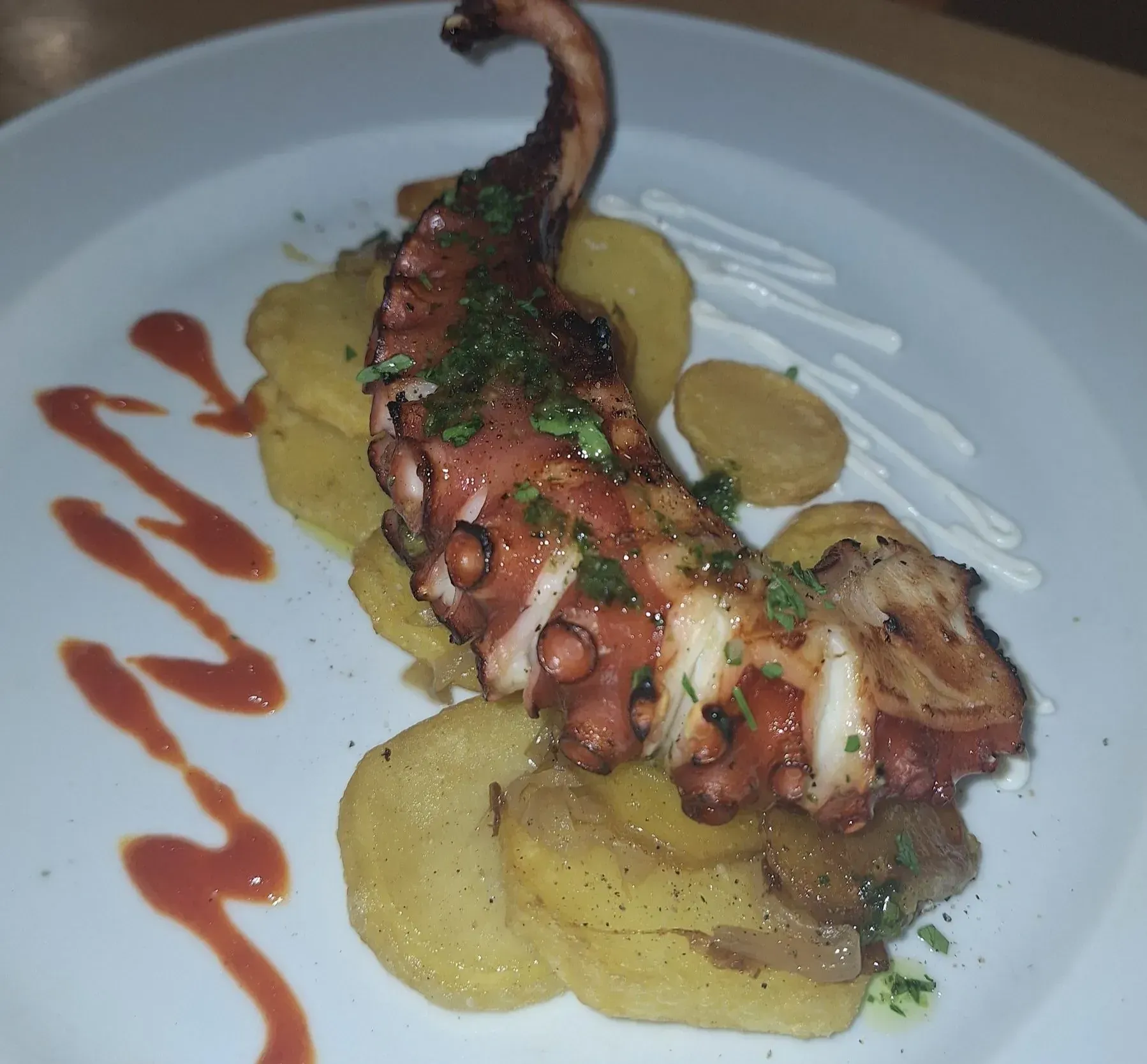 Tentáculo de pulpo a la parrilla sobre rodajas de patata frita, aderezado con salsas en un plato blanco.
