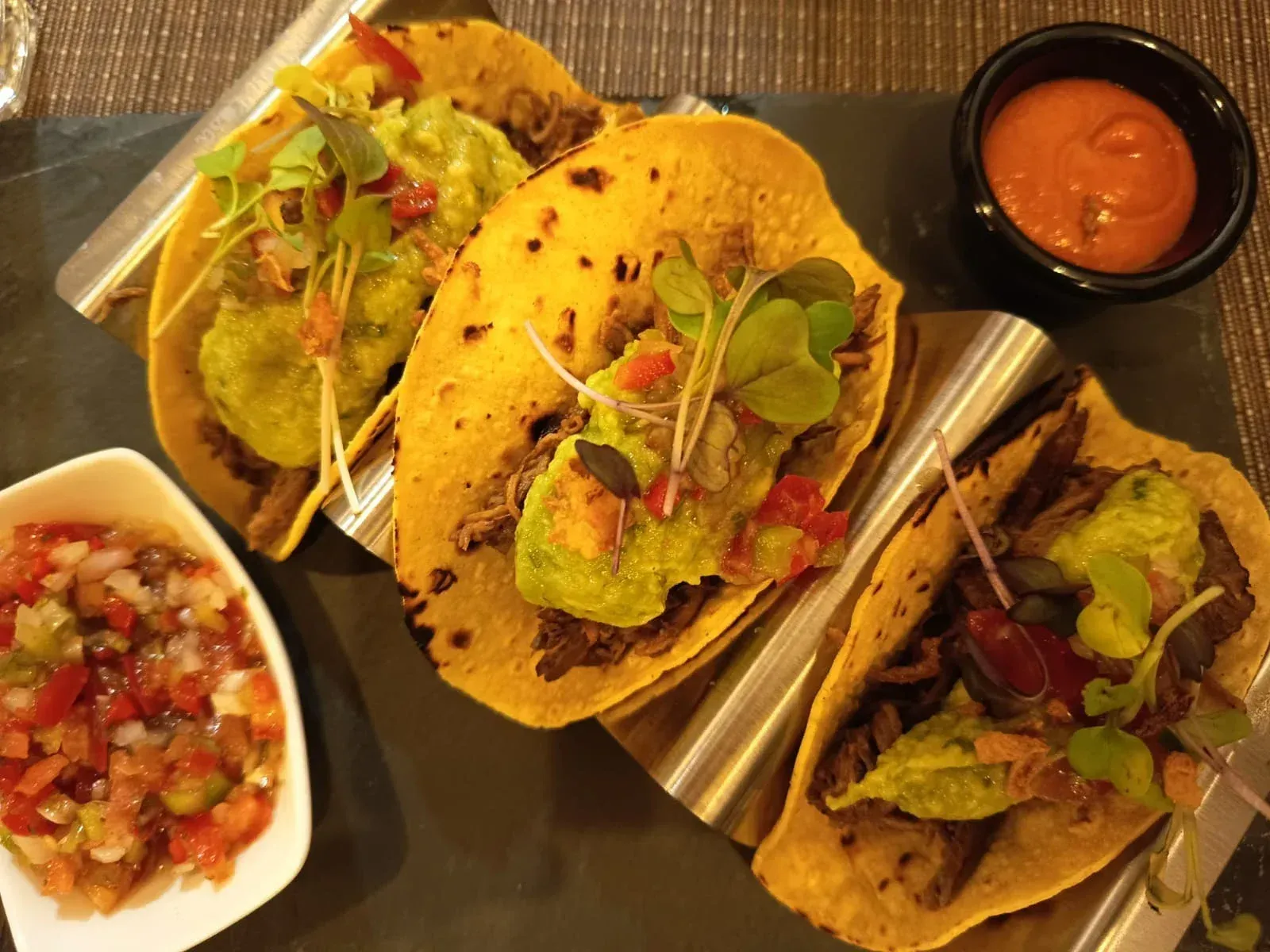 Tres tacos sobre una superficie oscura, cubiertos con guacamole y microvegetales, servidos con salsa y un tazón pequeño de salsa.
