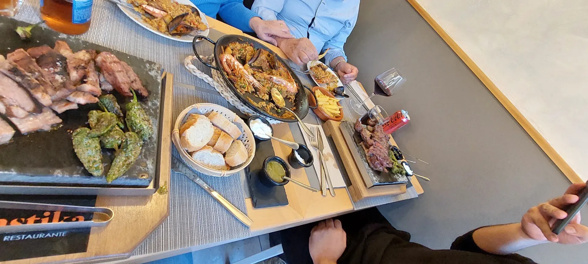 Vista aérea de una mesa con diversos platos, incluyendo filete, paella y pimientos. La gente está sentada alrededor de la mesa.