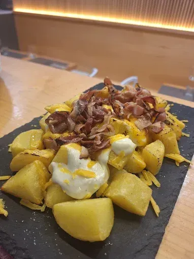Gajos de papa fritos dorados con queso, crema agria y trocitos de tocino sobre una pizarra negra.