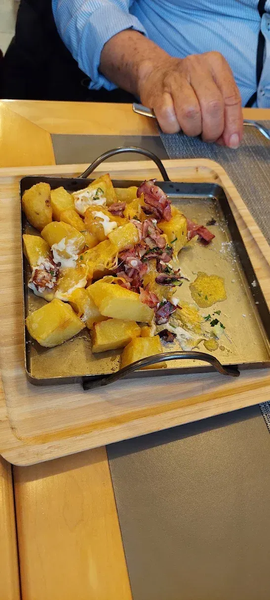 Un plato de patatas con aderezos reposa sobre una bandeja de madera en una mesa de restaurante. Se ven la mano y el brazo de una persona.