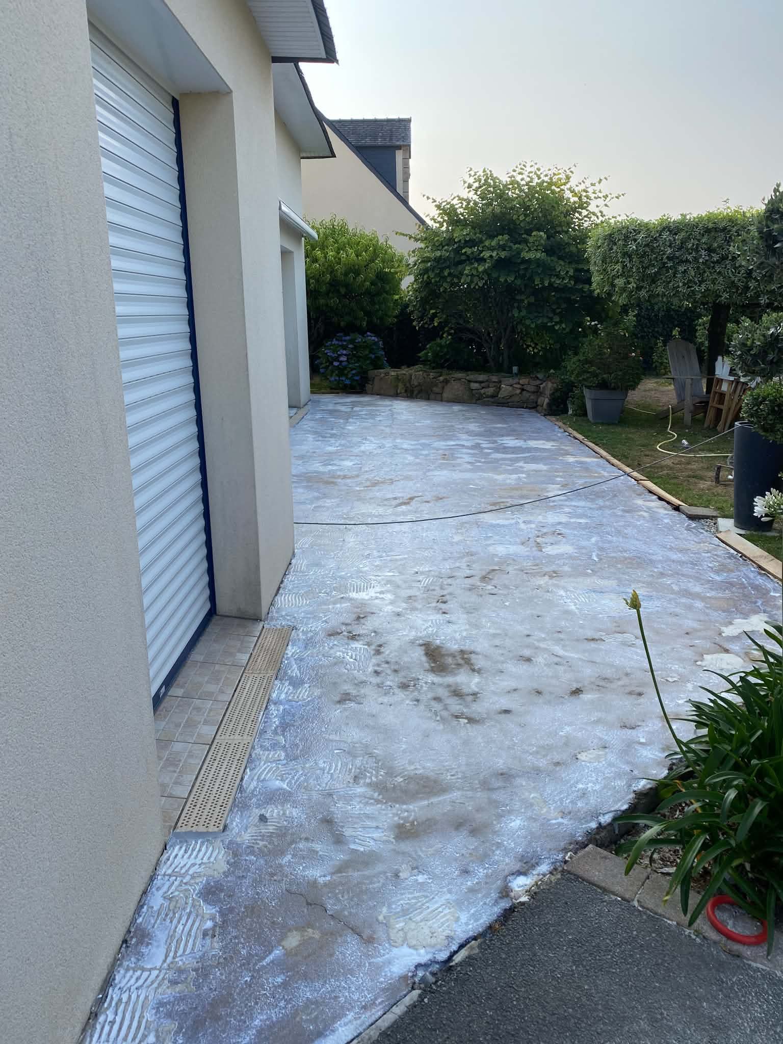 Allée en béton longeant une maison, bordée de buissons et d'arbres.