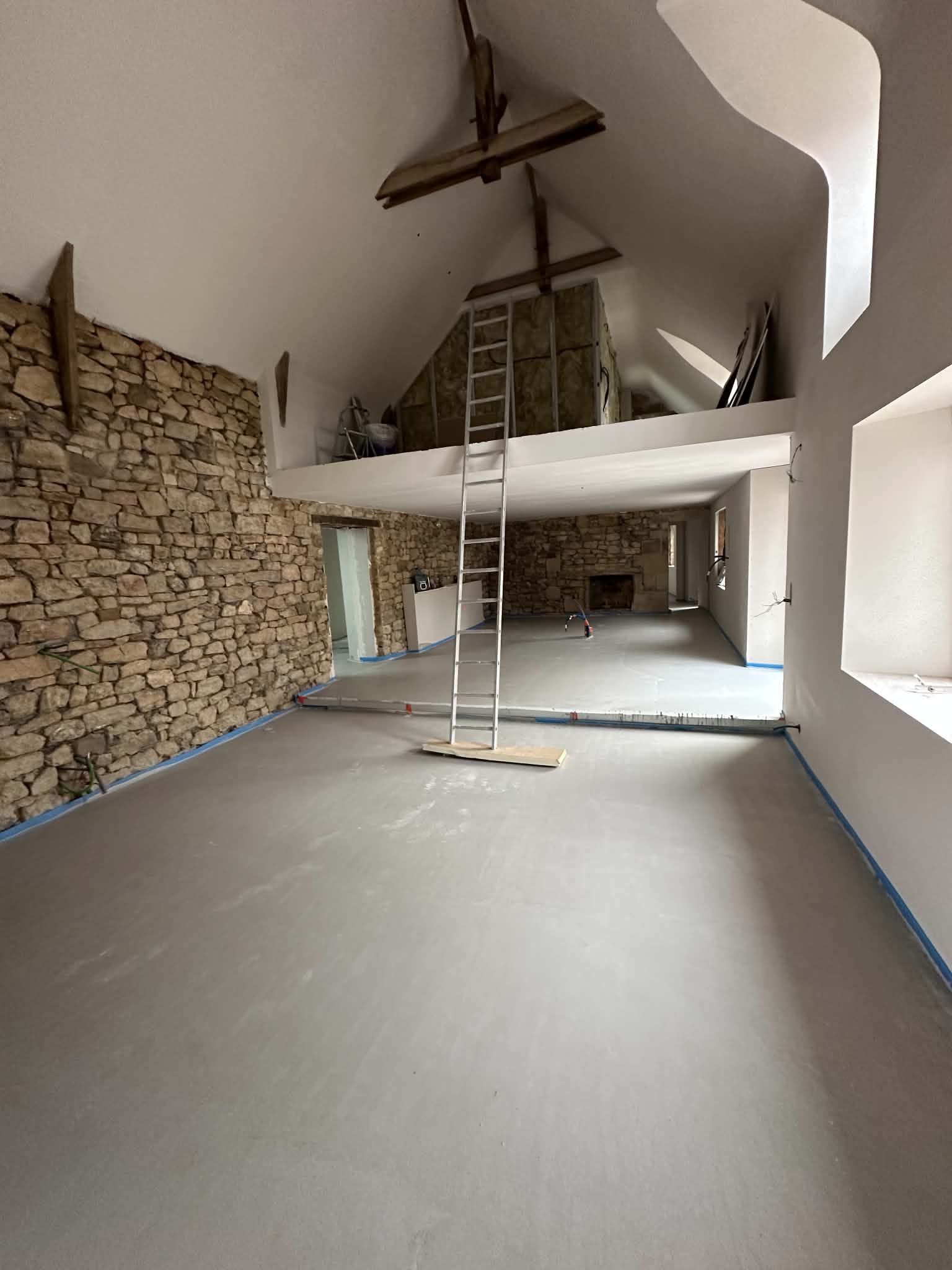 Pièce vide en rénovation, avec un mur en pierre, un haut plafond voûté et des échelles sur un sol.