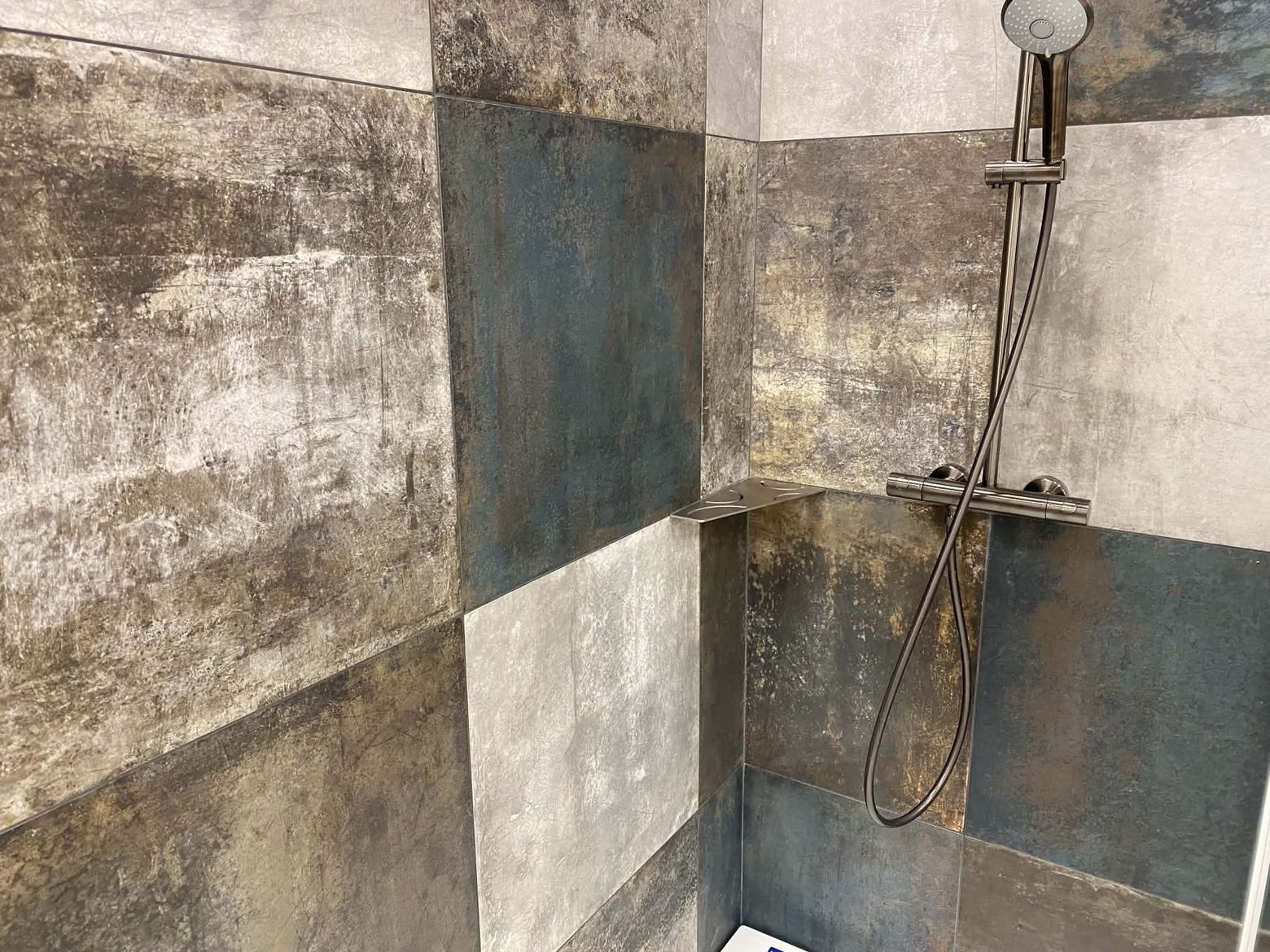 Coin de douche carrelé avec pommeau de douche mural et flexible, laissant apparaître des carreaux tachés.