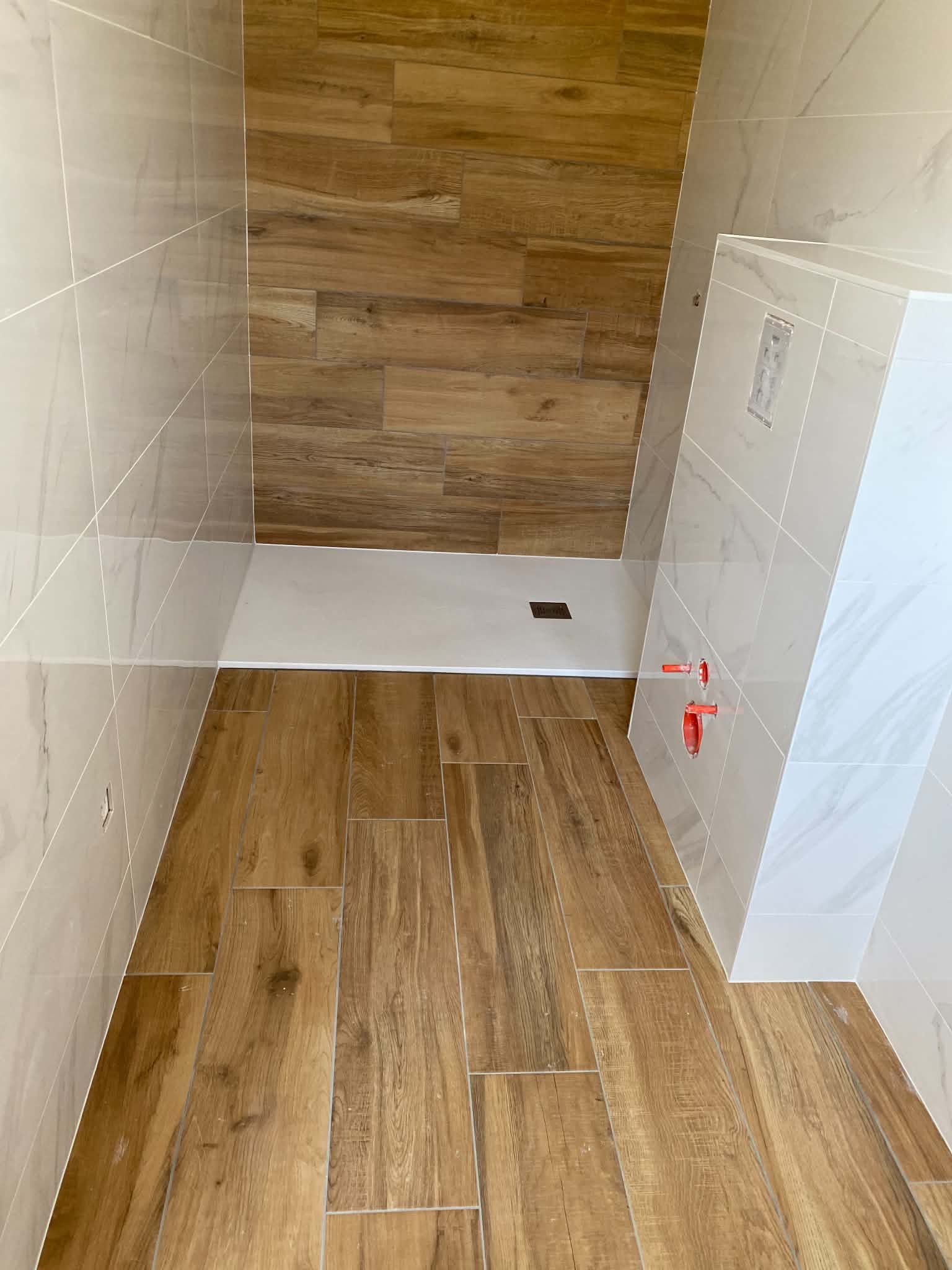 Salle de bains vide avec sol carrelé imitation bois, murs et un espace douche/baignoire au fond.