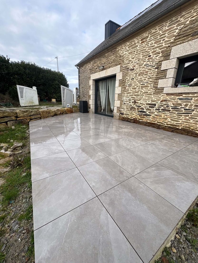 Terrasse en pierre humide jouxtant une maison en pierre avec portes vitrées et fenêtre, vue en angle.