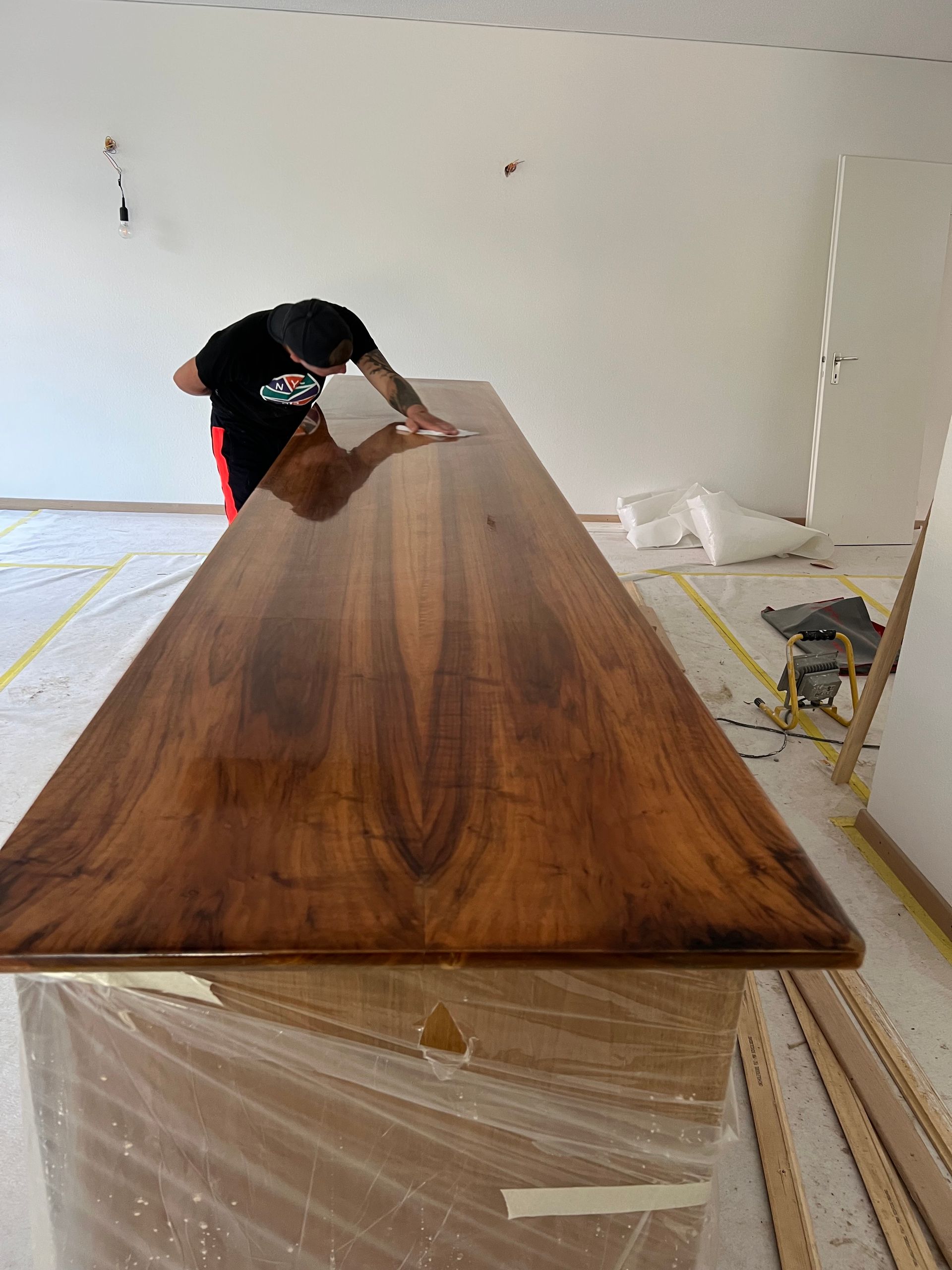 Ein Mann poliert einen langen Holztisch. Der Tisch hat eine markante, braune Maserung und steht in einem hellen, weißen Raum.