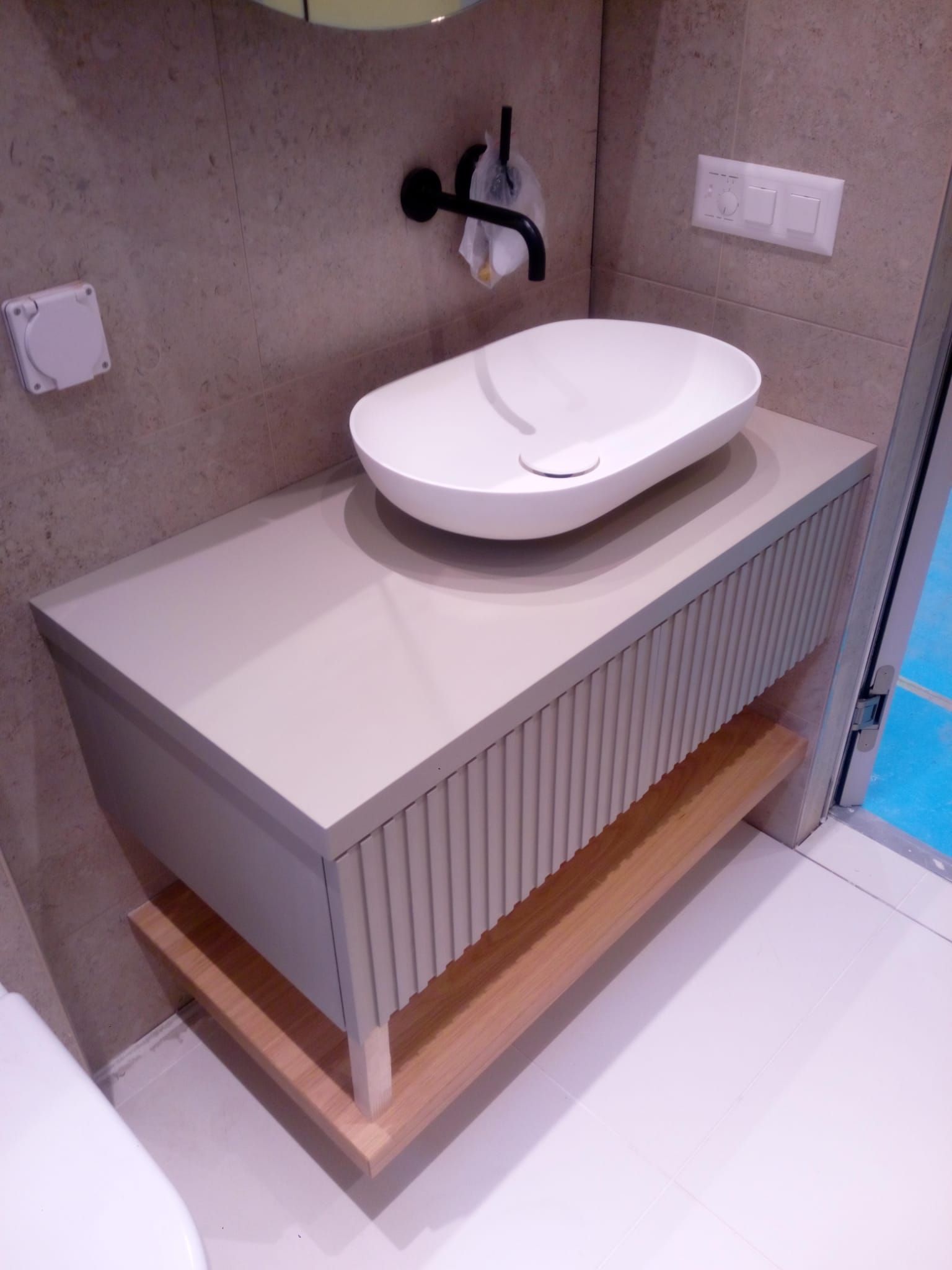 Badezimmerwaschtisch mit einem weißen ovalen Waschbecken auf einem grauen Schrank mit einem Holzregal und hellen Wänden.