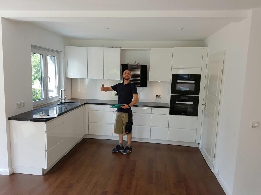 Ein Mann in einer frisch renovierten, weißen Küche zeigt den Daumen nach oben. Die Küche ist mit dunklen Arbeitsplatten, Holzböden und Einbaugeräten ausgestattet.